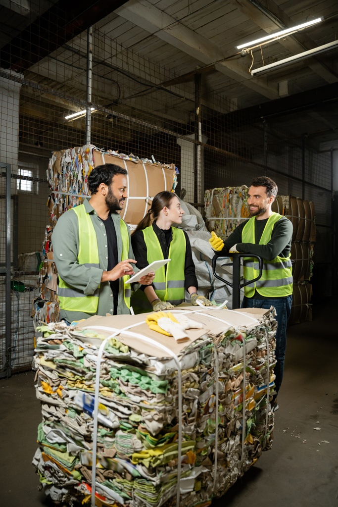 Cheerful sorter in protective vest and gloves talking to interracial colleagues with digital tablet and waste paper on hand pallet truck in waste disposal station, garbage recycling concept - Photo, Image