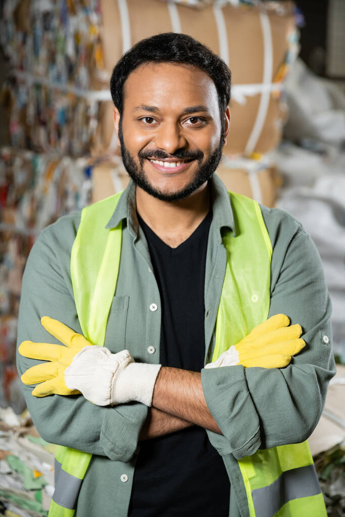 Cheerful indian worker in reflective vest and gloves looking at camera and crossing arms while standing near waste paper on blurred background in waste disposal station, garbage recycling concept - Photo, Image