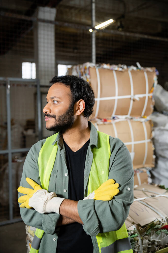 廃棄物処理場の紙の近くに立っている間、高い視認性のベストと手袋を離れて見て、腕を横断ひげそりインドの労働者、ゴミの分類とリサイクルの概念 - 写真・画像