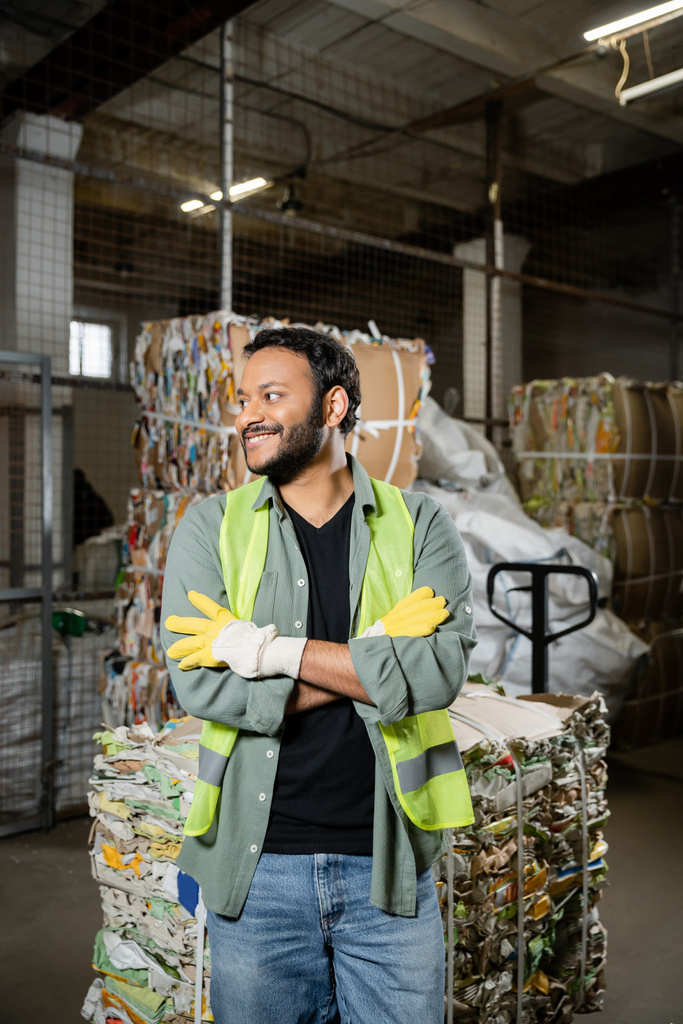 Positive indian sorter in protective gloves and vest crossing arms while standing near waste paper on hand pallet truck in blurred waste disposal station, garbage sorting and recycling concept - Photo, Image
