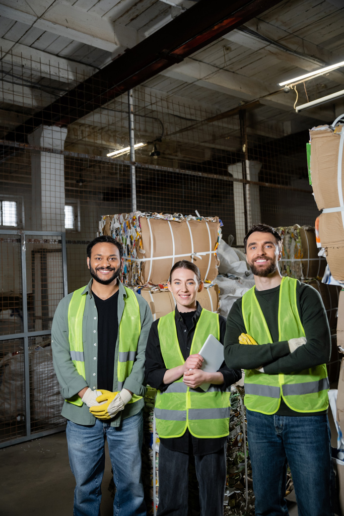 Smiling interracial sorters in protective vests holding digital tablet and looking at camera near waste paper in waste disposal station, garbage sorting and recycling concept - Photo, Image