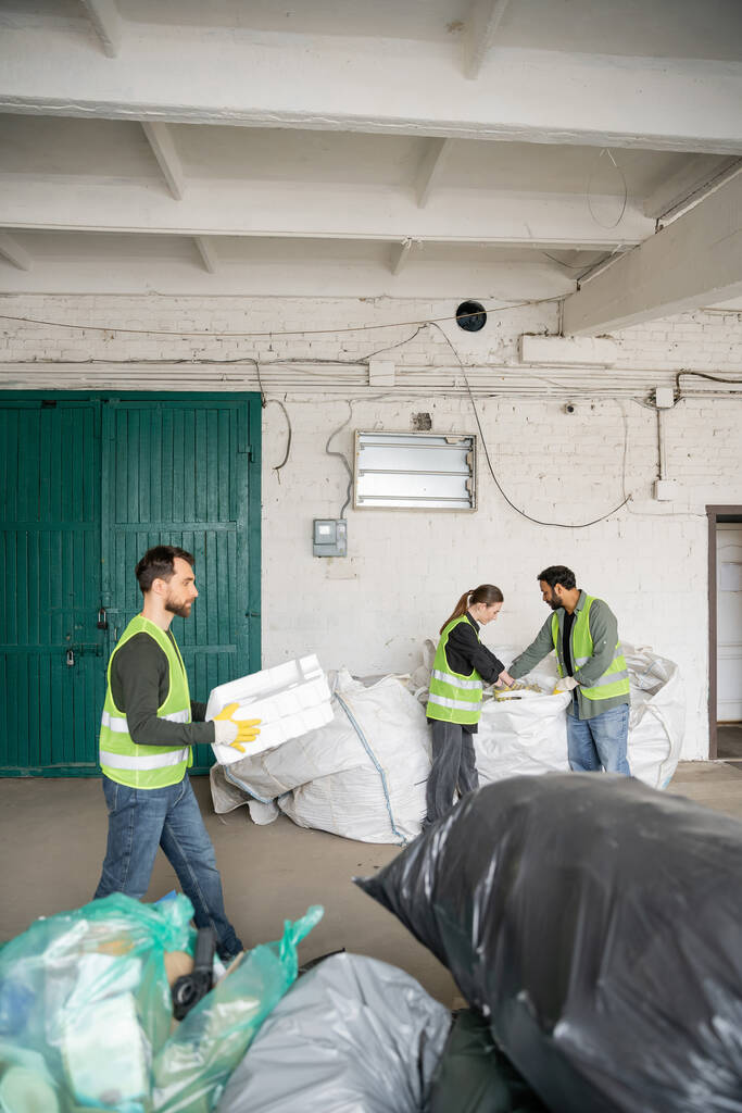Trabalhador do sexo masculino em colete de alta visibilidade e luvas transportando espuma de plástico, enquanto colegas inter-raciais trabalhando com sacos na estação de descarte de resíduos, triagem de lixo e conceito de reciclagem - Foto, Imagem