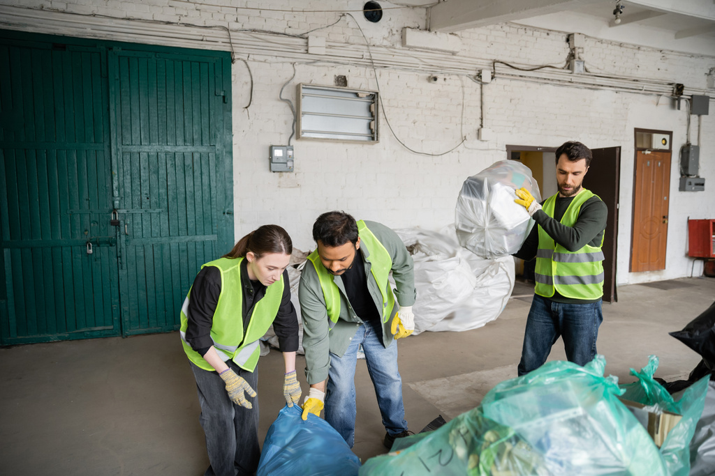 Monietniset työntekijät suojaavissa liiveissä ja käsineissä, jotka työskentelevät muovipussien kanssa lähellä kollegaa jätteenkäsittelyasemalla, jätteiden lajittelu ja kierrätys - Valokuva, kuva