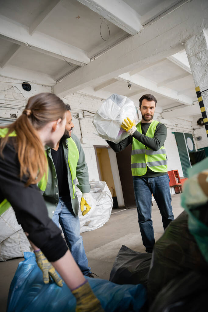 Εργαζόμενος με προστατευτικό γιλέκο και γάντι κρατώντας πλαστική σακούλα με σκουπίδια κοντά σε θολή διαφυλετικούς συναδέλφους που εργάζονται στο σταθμό διάθεσης αποβλήτων, διαλογή και ανακύκλωση έννοια - Φωτογραφία, εικόνα