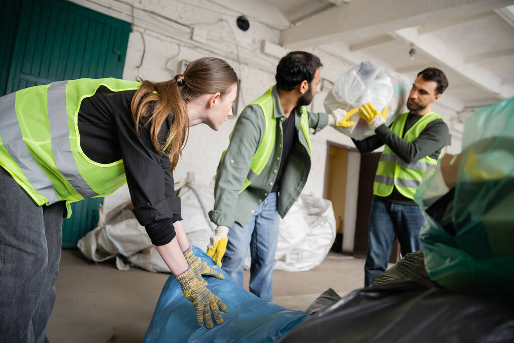 Молодая работница в защитном жилете и перчатках с пластиковым пакетом рядом с размытыми многонациональными коллегами на станции по утилизации отходов, концепция сортировки и переработки мусора - Фото, изображение