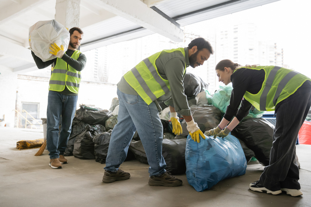 Uśmiechnięta pracownica w kamizelce i rękawiczkach, wkładająca plastikową torbę ze śmieciami w pobliżu wielonarodowych kolegów z zakładu utylizacji odpadów, koncepcja sortowania i recyklingu odpadów - Zdjęcie, obraz