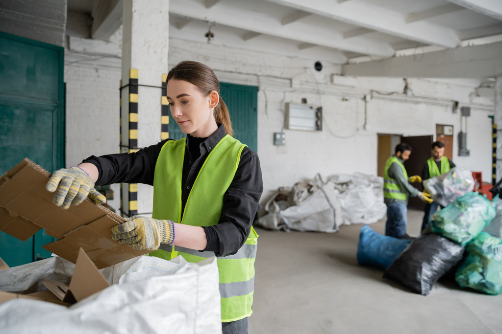Νεαρή εργαζόμενη με προστατευτικό γιλέκο και γάντια που κρατά χαρτόνι κοντά σε σάκους και θολή συναδέλφους που εργάζονται σε σταθμό διάθεσης αποβλήτων, διαλογή και ανακύκλωση - Φωτογραφία, εικόνα
