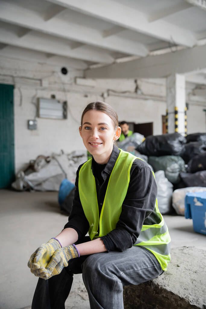 Positiivinen nuori työntekijä suojaavissa liiveissä ja käsineissä katselee kameraa, kun lepää lähellä hämärtyneitä muovipusseja, joissa on roskia taustalla roskien lajittelukeskuksessa, kierrätyskonsepti - Valokuva, kuva