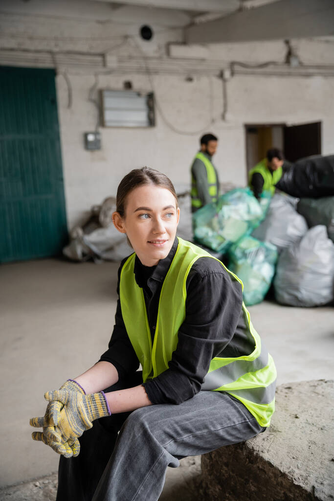 Positive young sorter in high visibility vest and gloves looking away while relaxing near blurred colleagues and plastic bags at background in garbage sorting center, recycling concept - Photo, Image