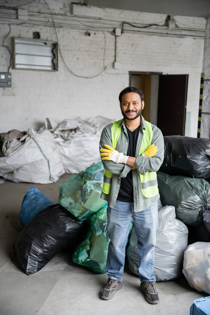 Travailleur indien joyeux et barbu en gilet de protection et gants croisant les bras et regardant la caméra tout en se tenant près des sacs en plastique dans le centre de tri des ordures, concept de recyclage - Photo, image