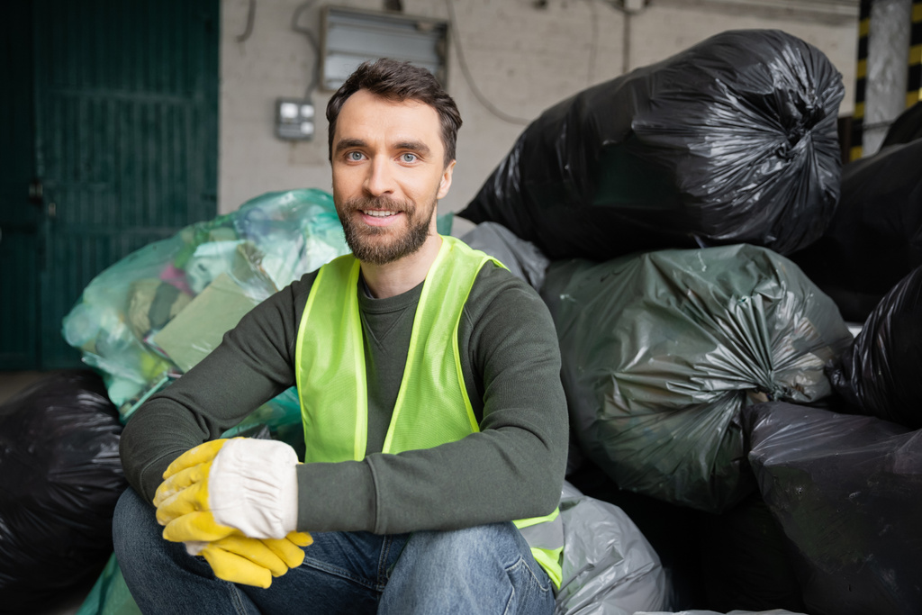 Trabajador sonriente y barbudo en chaleco de seguridad y guantes mirando a la cámara mientras está sentado en bolsas de plástico con basura en el centro de clasificación de basura borrosa, concepto de reciclaje - Foto, Imagen