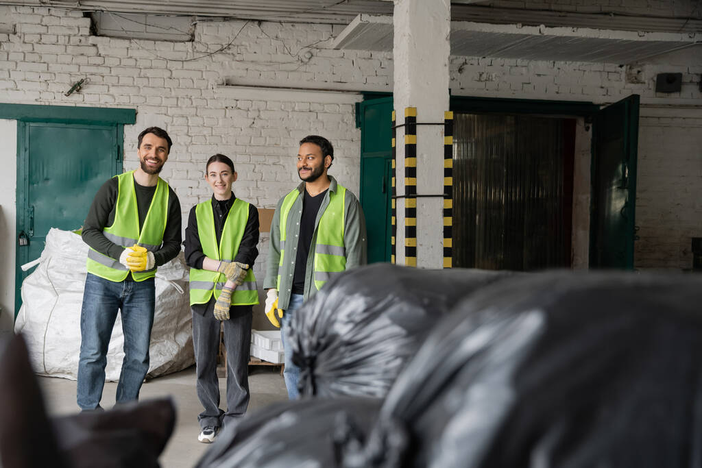 Trabajadores multiétnicos positivos en chalecos fluorescentes y guantes protectores mirando a la cámara mientras están de pie cerca de bolsas de plástico borrosas en el centro de clasificación de basura, concepto de reciclaje - Foto, imagen