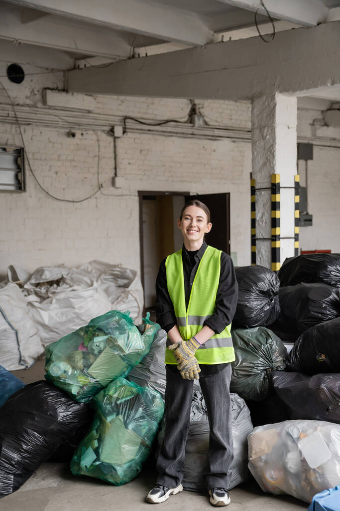 Hymyilevä naispuolinen työntekijä suojakäsineissä ja liiveissä katselee kameraa seisoessaan muovipussien lähellä roskien lajittelukeskuksessa, kierrätyskonsepti - Valokuva, kuva