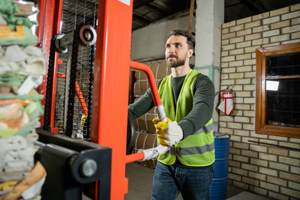 Bearded worker in fluorescent vest and gloves using hand pallet truck with waste paper while working in blurred garbage sorting center at background, recycling concept - Photo, Image