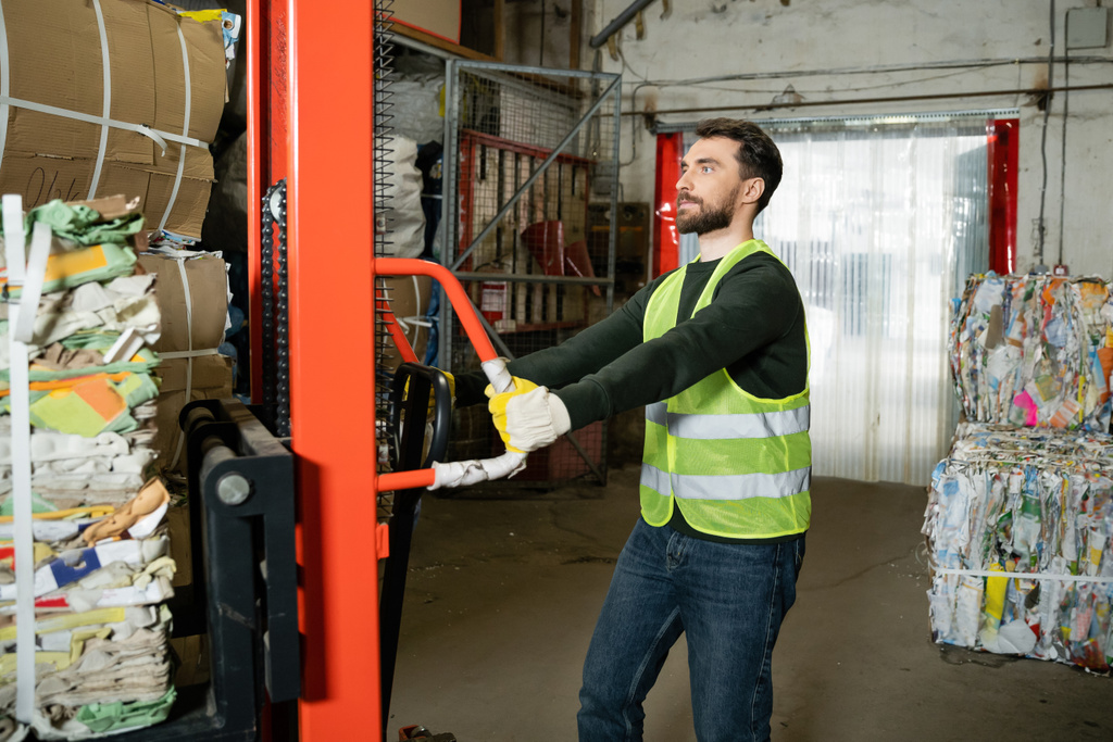 Bearded sorter in protective glove and vest using hand pallet truck with waste paper while working in blurred garbage sorting center at background, recycling concept - Photo, Image