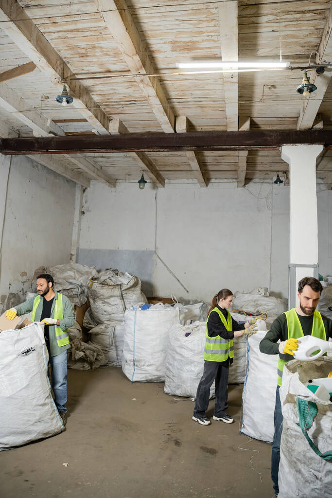 Interraciale werknemers in goed zichtbare vesten en handschoenen die afval in zakken sorteren terwijl zij samenwerken in afvalverwijderingsstation, afvalsorteer- en recyclingconcept - Foto, afbeelding
