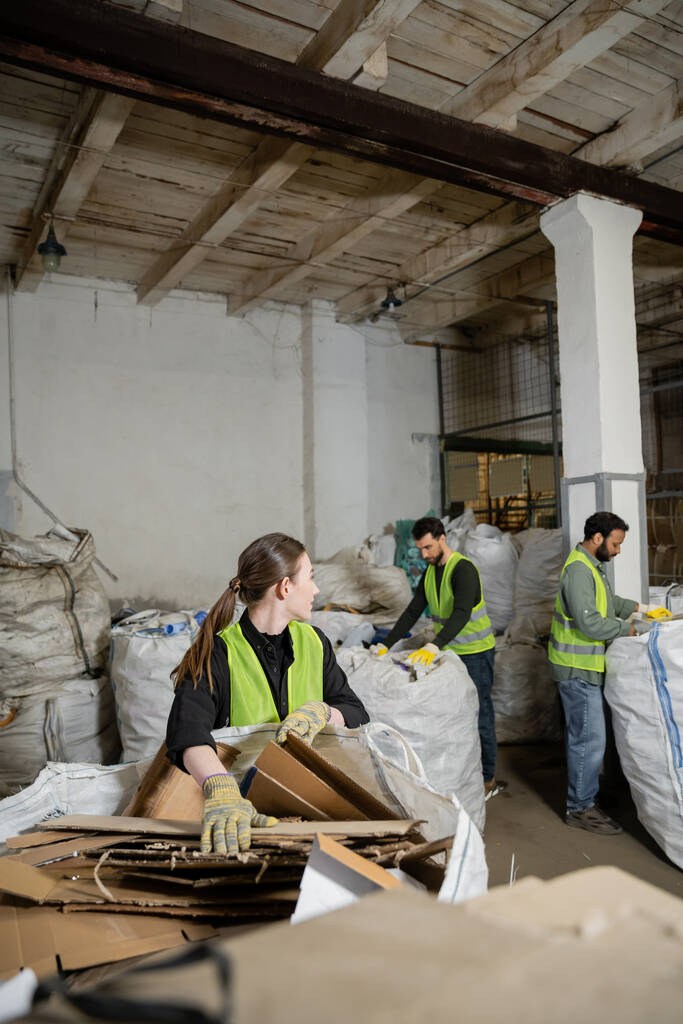 Νεαρή εργαζόμενη με προστατευτικά γάντια και γιλέκο που κοιτάζει πολυεθνικούς συναδέλφους ενώ βάζει χαρτόνι σε σάκους στο σταθμό διάθεσης αποβλήτων, έννοια διαλογής απορριμμάτων και ανακύκλωσης - Φωτογραφία, εικόνα