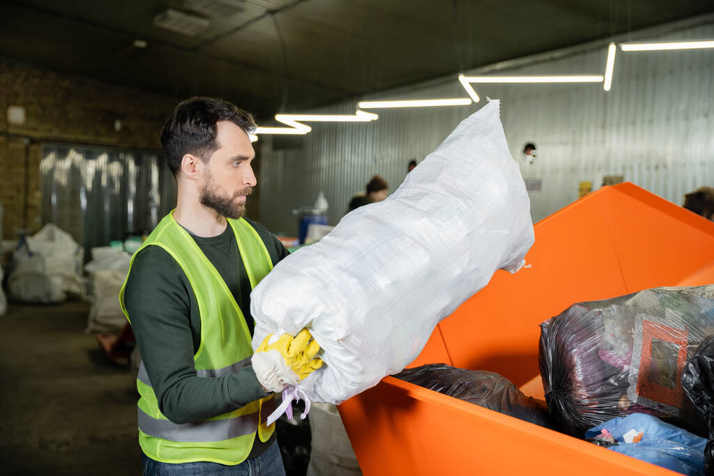 Zijaanzicht van bebaarde sorteerder in handschoen en beschermvest die zak met afval in doos in wazig afvalverwijderingsstation, afvalsorteer- en recyclingconcept stopt - Foto, afbeelding