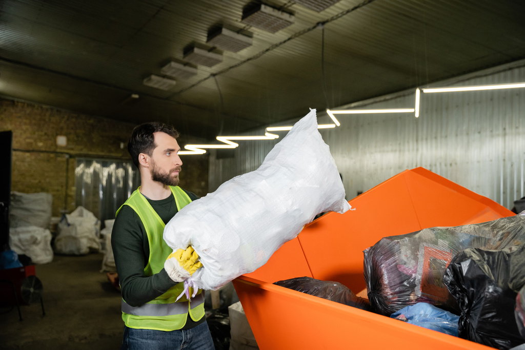 Πλευρική άποψη του ανθρώπου σε γιλέκο υψηλής ορατότητας και γάντι βάζοντας σάκο με σκουπίδια σε δοχείο, ενώ εργάζονται σε σταθμό διάθεσης αποβλήτων, διαλογή και ανακύκλωση έννοια - Φωτογραφία, εικόνα