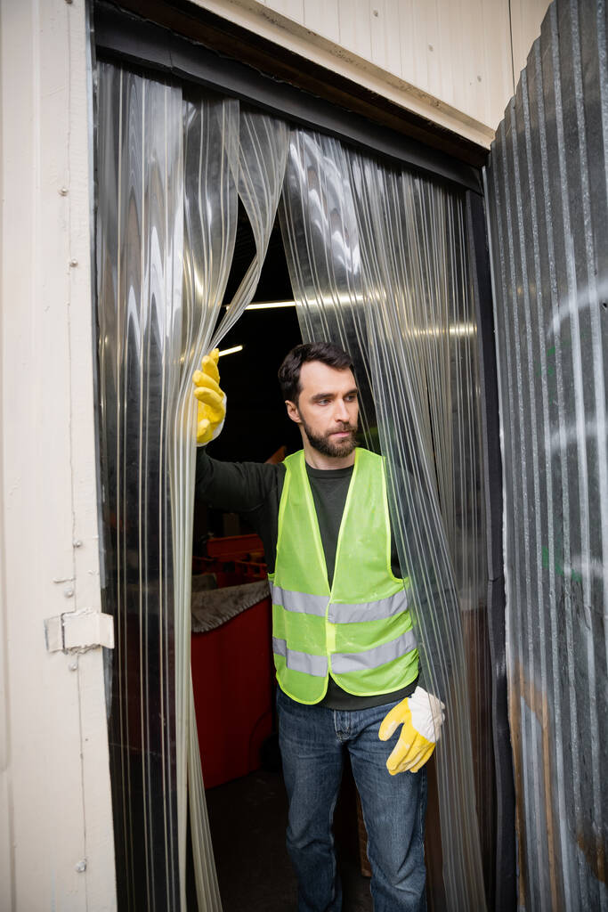 Γενειοφόρος εργάτης με γιλέκο υψηλής ορατότητας και γάντια που στέκονται κοντά στην είσοδο του σταθμού διάθεσης αποβλήτων, έννοια διαλογής και ανακύκλωσης απορριμμάτων - Φωτογραφία, εικόνα