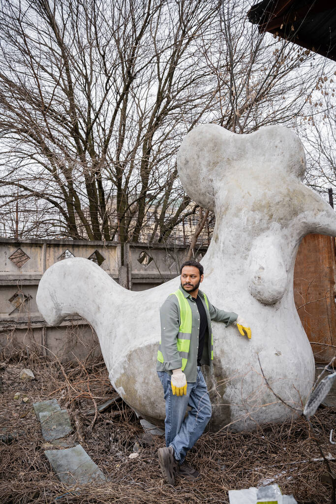 Indischer Arbeiter in Warnweste und Handschuhen schaut weg, während er in der Nähe einer Betonskulptur in einer Außendeponie steht, Müllsortier- und Recyclingkonzept - Foto, Bild