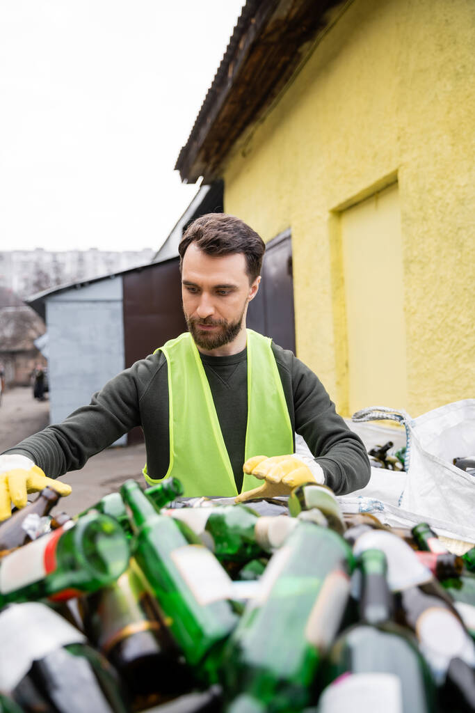 Bearded male worker in protective vest and gloves standing near blurred glass trash in outdoor waste disposal station, garbage sorting and recycling concept - Photo, Image