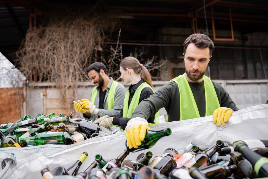 Bärtige Arbeiter in Schutzhandschuhen und Weste stecken Glasflasche in Sack neben verschwommenen Kollegen in Müllentsorgungsstation, Müllsortier- und Recyclingkonzept - Foto, Bild
