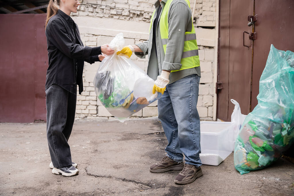 Gestroopt zicht op glimlachende vrijwilliger geven plastic zak met afval aan de werknemer in beschermende vest en handschoenen in de buurt van afval buiten in afvalverwerkingsstation, afval sorteren en recycling concept - Foto, afbeelding