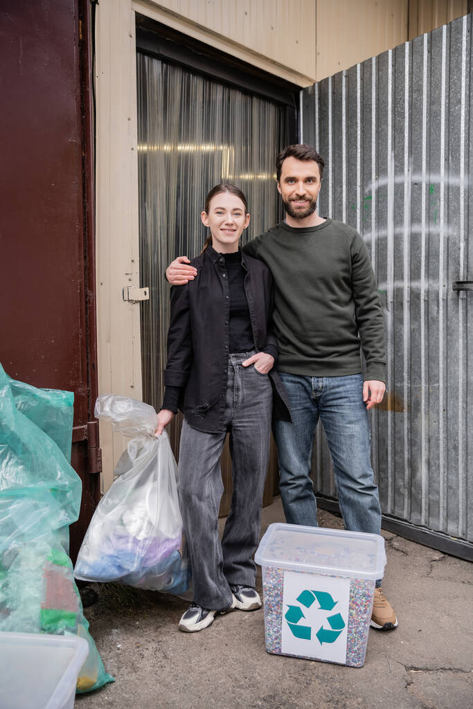 Hymyilevät vapaaehtoiset katselevat kameraa seistessään kyynelten roskat ja ovi jätteenkäsittelyaseman ulkona, roskien lajittelu ja kierrätys käsite - Valokuva, kuva