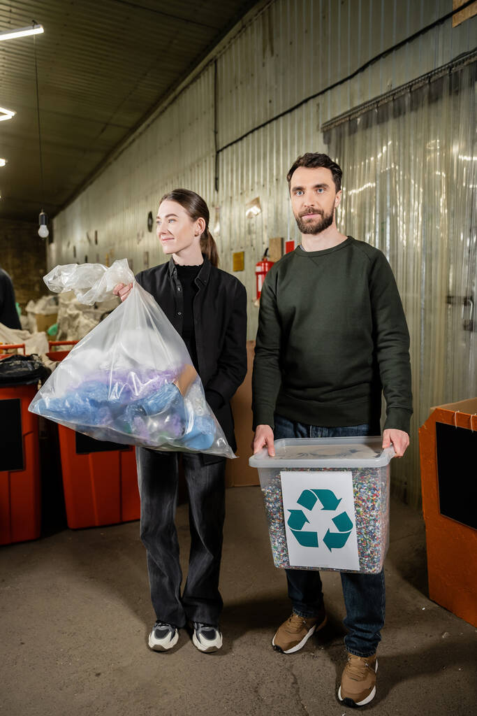 Позитивные волонтеры держат мусорный бак и мешок, стоя вместе в размытой станции утилизации отходов на заднем плане, сортировки и переработки мусора концепции - Фото, изображение