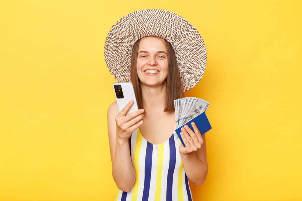 Усміхнена приваблива жінка турист використовує мобільний телефон, стоячи з квитками паспорт в руках, що виїжджають за кордон в смугастому плавальному костюмі, позує ізольовано на жовтому тлі
 - Фото, зображення
