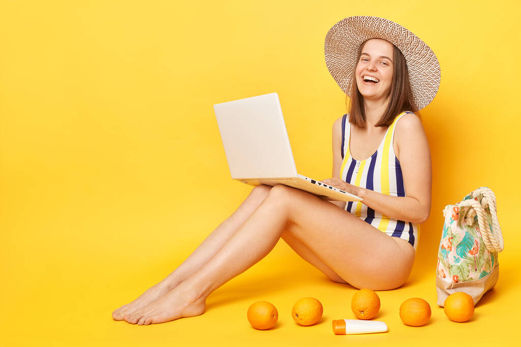 Onnellinen optimistinen nainen yllään raidallinen uimapuku ja hattu eristetty keltaisella taustalla työskentelevät kannettavan nauraa iloisesti tyhjä tila. - Valokuva, kuva