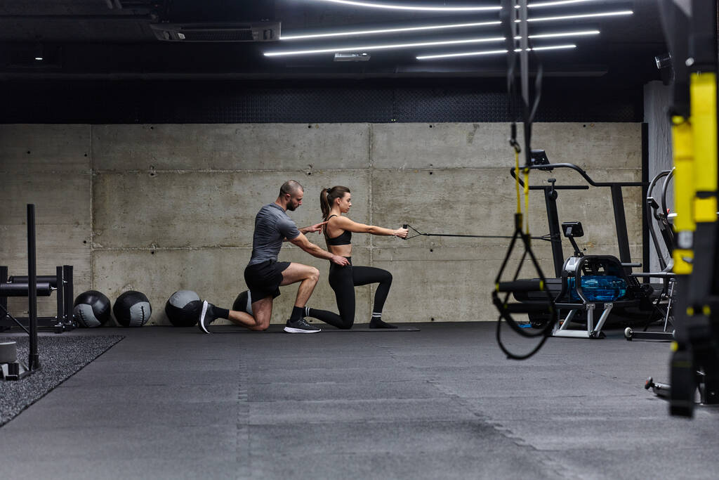 Muskularny mężczyzna pomagający sprawnej kobiecie w nowoczesnej siłowni, gdy wykonuje różne ćwiczenia na ciało i rozciąga mięśnie, pokazując swoje zaangażowanie w fitness i korzystając z pracy zespołowej i wsparcia. - Zdjęcie, obraz