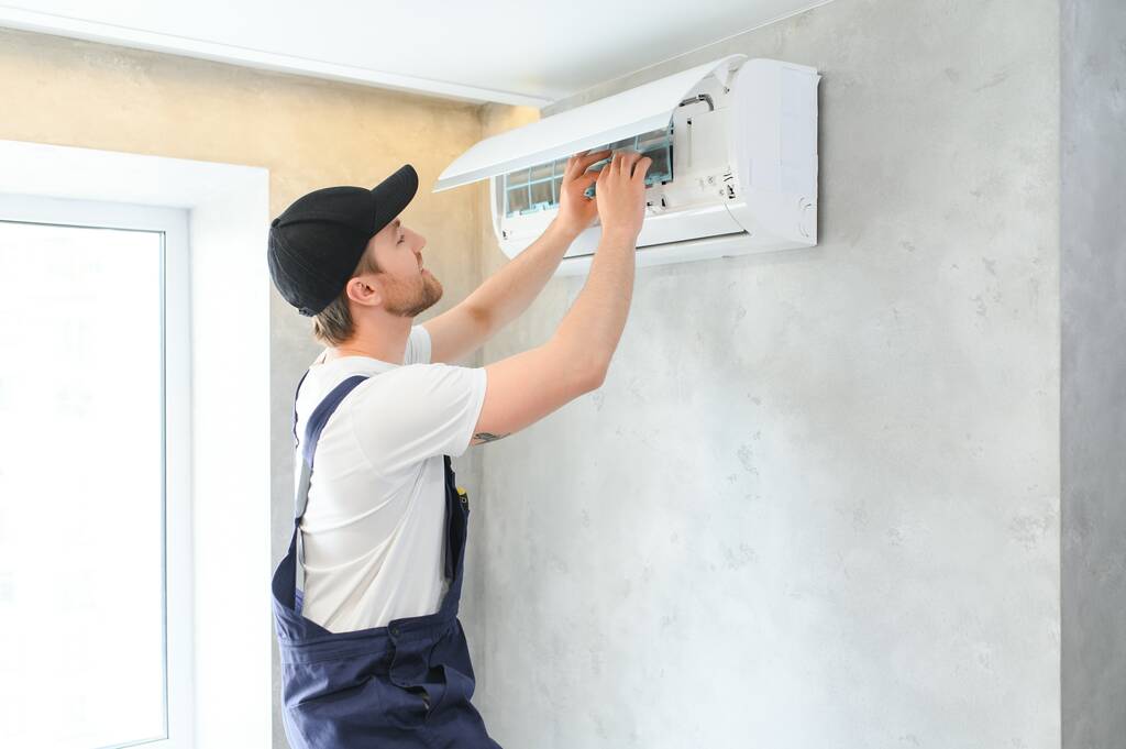Mosolygó fiatal férfi villanyszerelő javító kondicionáló az ügyfél otthonában. Férfi szerelő vagy szerelő javítás szűrők légkondicionáló készülék ügyfélház. - Fotó, kép