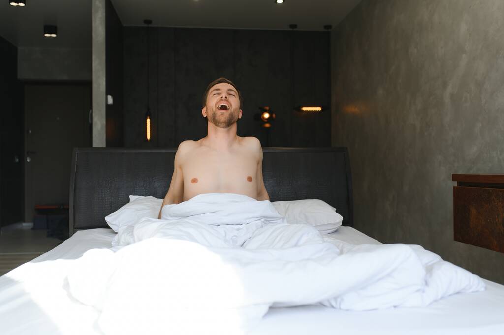 Νεαρός άνδρας ξυπνά και σηκώνεται με τα χέρια τεντωμένα στο λευκό κρεβάτι το πρωί με το φως του ήλιου μέσα από το παράθυρο. - Φωτογραφία, εικόνα