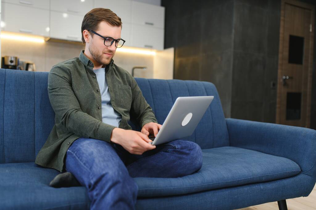 Ικανοποιημένος όμορφος νεαρός καυκάσιος άνδρας με casual stylish ρούχα, κάθεται στον καναπέ στο σπίτι στο άνετο σαλόνι, χρησιμοποιώντας ένα φορητό υπολογιστή, εργασίας ή μελέτης σε απευθείας σύνδεση, ονειρεύεται, χαμογελά. - Φωτογραφία, εικόνα
