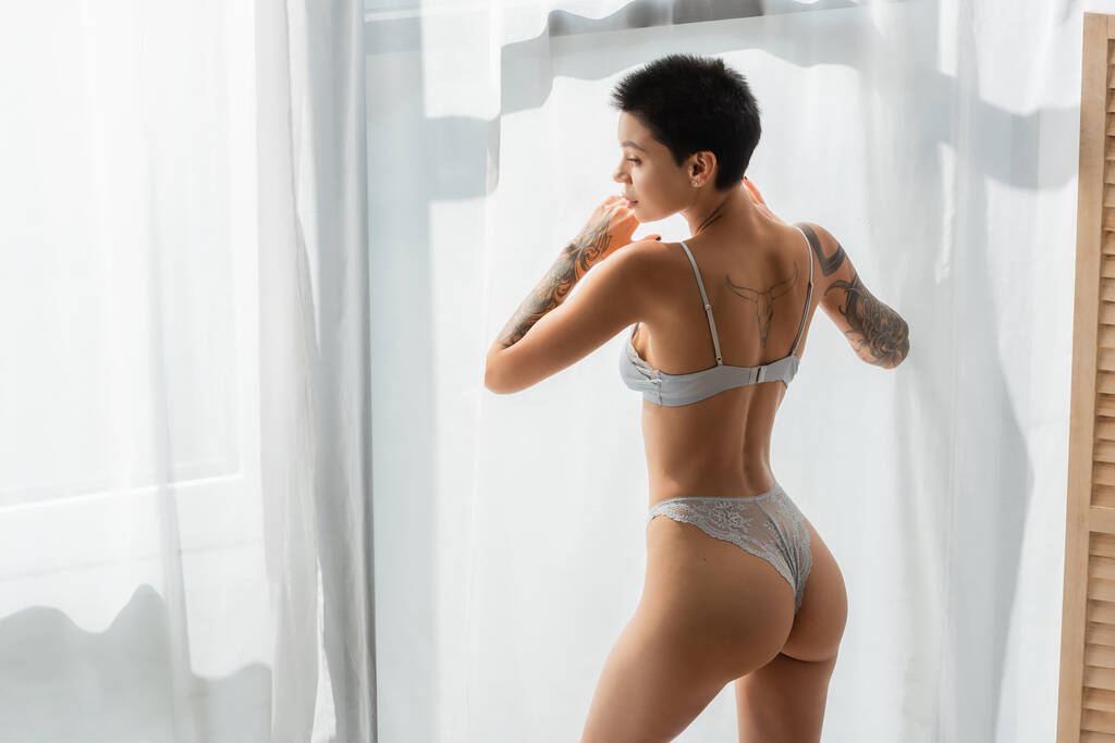 young and stunning woman with sexy tattooed body and short brunette hair wearing lace panties and bra while standing in natural light near white curtain in modern bedroom at home - Photo, Image