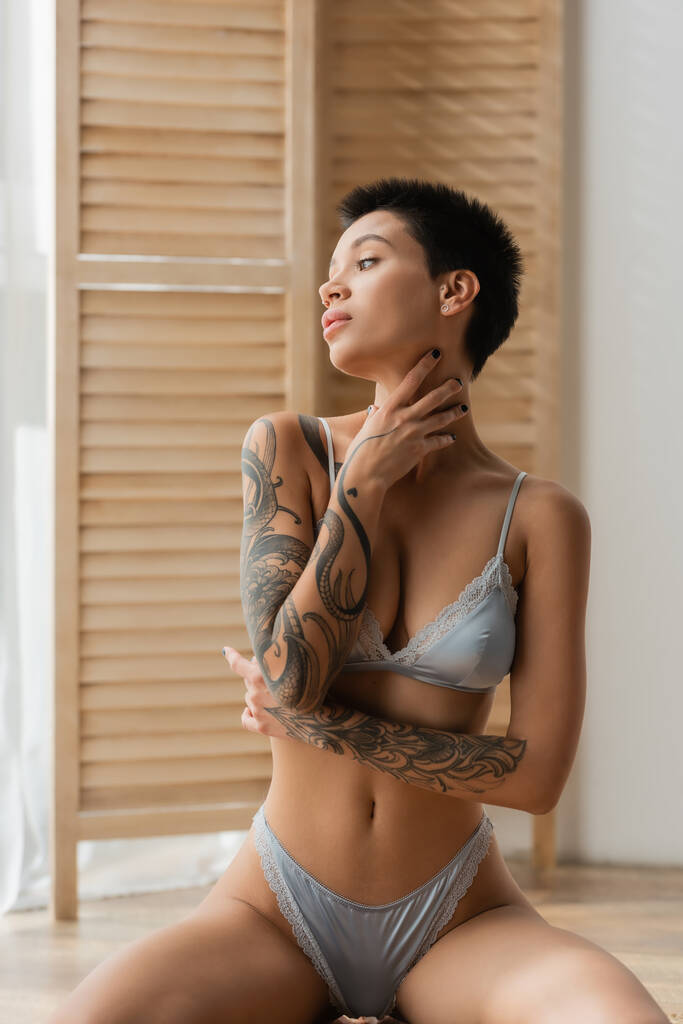 young and tempting woman with tattooed body sitting in seductive pose in grey silk lingerie, touching neck and looking away near room divider on blurred background in bedroom - Photo, Image