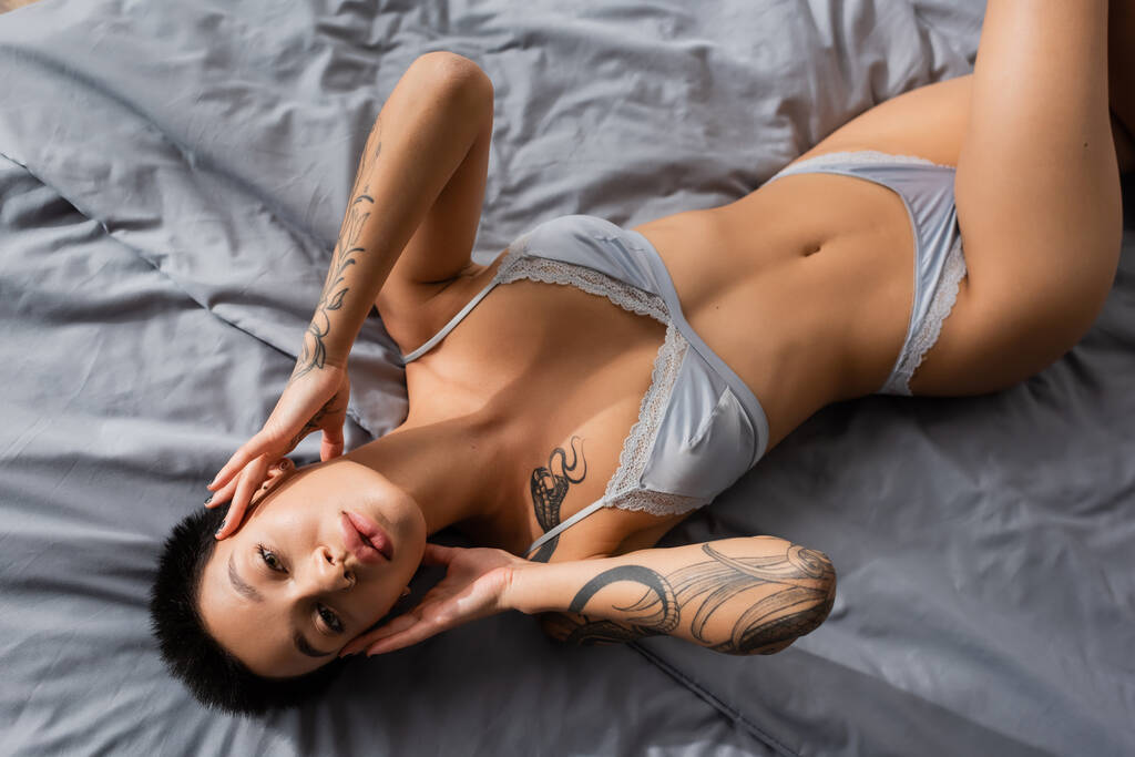 top view of sultry woman in lingerie, with sexy tattooed body and short brunette hair touching face and looking at camera on grey bedding in bedroom at home - Photo, Image