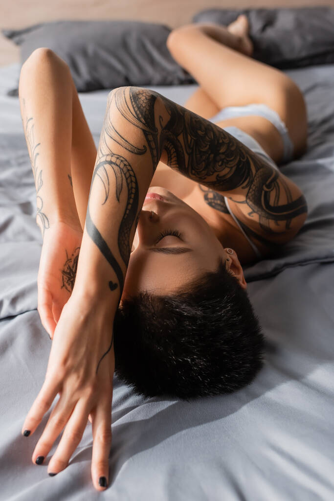young and passionate woman with closed eyes and sexy tattooed body posing in lingerie on grey bedding near pillows on blurred background in modern bedroom at home - Photo, Image