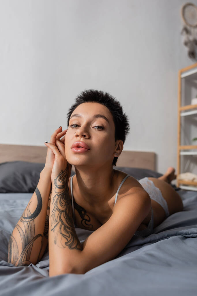 mujer encantadora en lencería, con el cuerpo sexy tatuado y pelo corto morena puesta en la ropa de cama gris y mirando a la cámara cerca de rack y almohadas sobre fondo borroso - Foto, Imagen