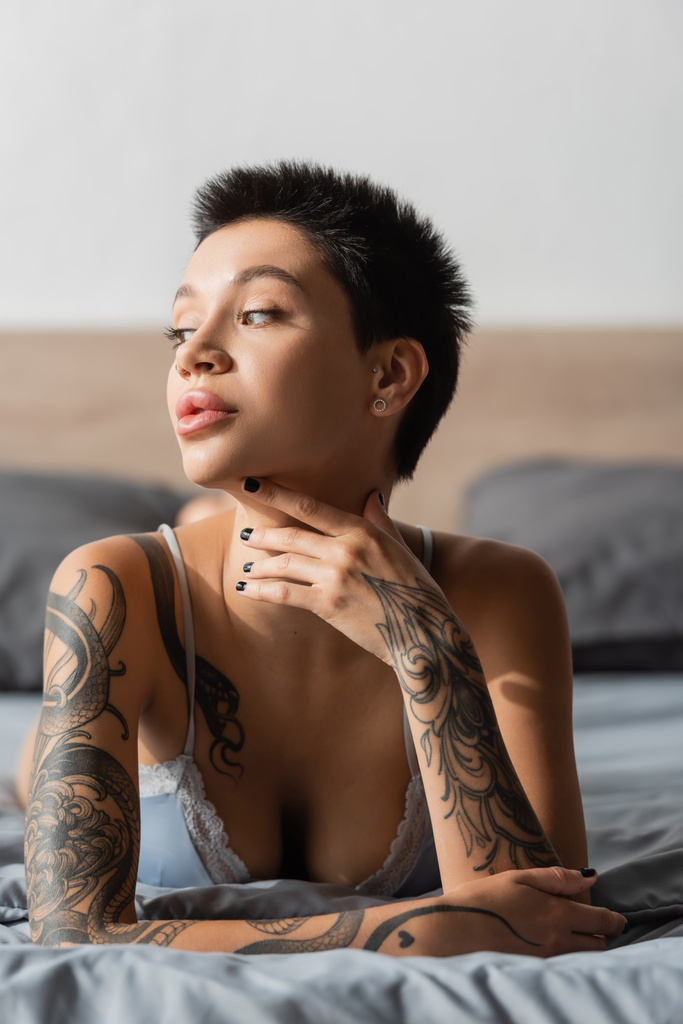 stunning woman with short brunette hair, tattooed body and sexy breast holding hand near neck while laying in bra on grey bedding and looking away on blurred background, boudoir photography - Photo, Image