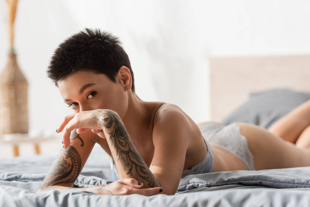 joven mujer intrigante y sexy con el pelo corto morena y el cuerpo tatuado mirando a la cámara mientras está acostado en ropa de cama gris en el sujetador y cogido de la mano cerca de la cara, fotografía boudoir  - Foto, Imagen