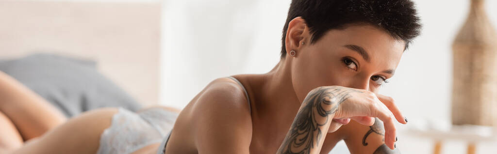 mulher jovem e sedutora com corpo tatuado sexy e cabelo curto morena olhando para a câmera e segurando a mão perto do rosto enquanto estava deitado no quarto moderno em casa, fotografia boudoir, banner - Foto, Imagem