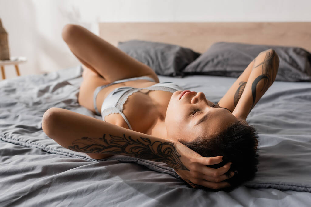 молодая, сексуальная и желанная женщина с закрытыми глазами и татуированным телом, прикасаясь к коротким волосам брюнеток, лежа рядом с подушками на размытом фоне в спальне, будуарная фотография - Фото, изображение