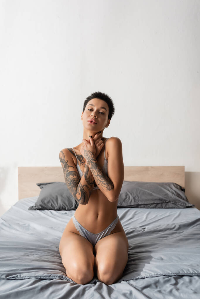 young appealing woman with short brunette hair and sexy tattooed body sitting on grey bedding in lingerie and looking at camera near pillows in modern bedroom, boudoir photography - Photo, Image