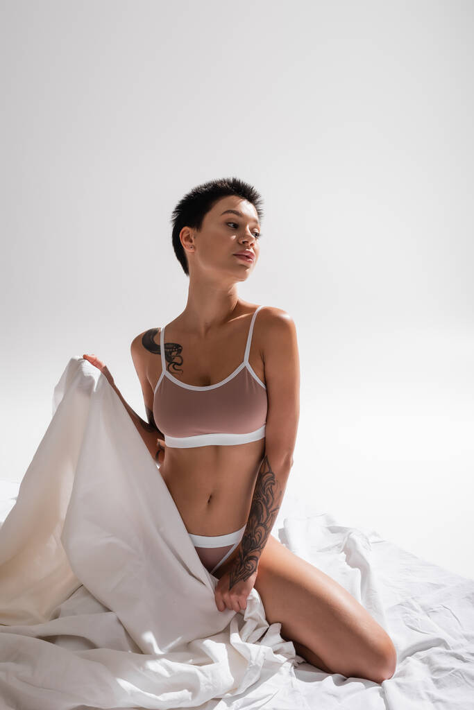young, tattooed and passionate woman in beige lingerie, with sexy body and short brunette hair holding white bed sheet and looking away in studio on grey background, erotic photography - Photo, Image