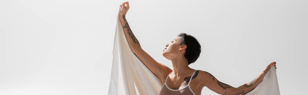 mujer joven y atractiva en sujetador beige, con cuerpo tatuado sexy y pelo corto morena sosteniendo sábana blanca mientras posa sobre fondo gris en el estudio, pancarta - Foto, imagen