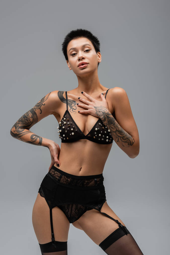 provocative woman with sexy tattooed body and short brunette hair, in bra with pearl beads, black panties, garter belt and stockings posing with hand on hip and looking at camera on grey background - Photo, Image