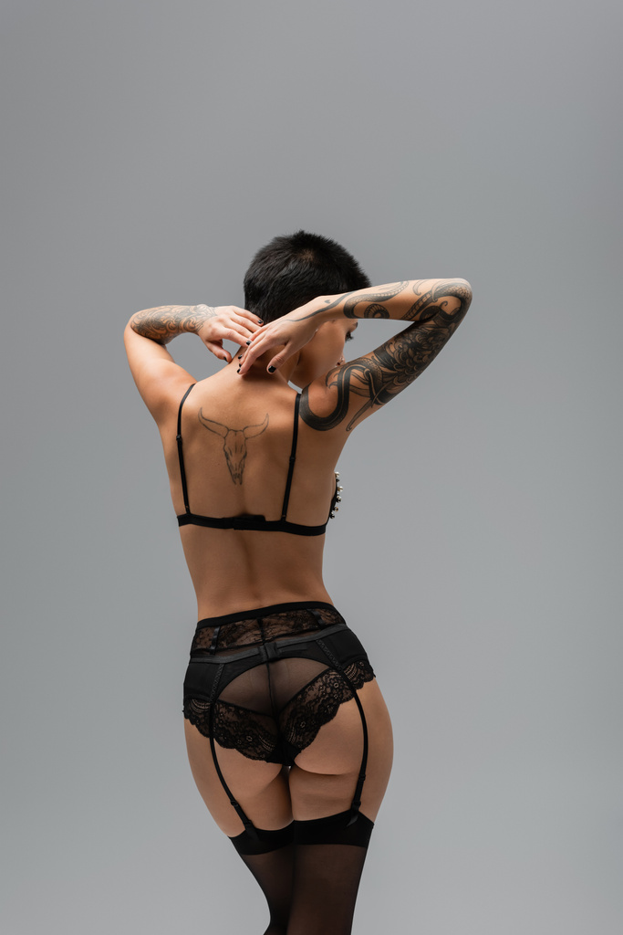zadní pohled neodolatelné ženy s tetovaným tělem a sexy hýždě nosí černou podprsenku, krajkové kalhotky, podvazkový pás a černé punčochy, zatímco stojí s rukama za krkem na šedém pozadí - Fotografie, Obrázek